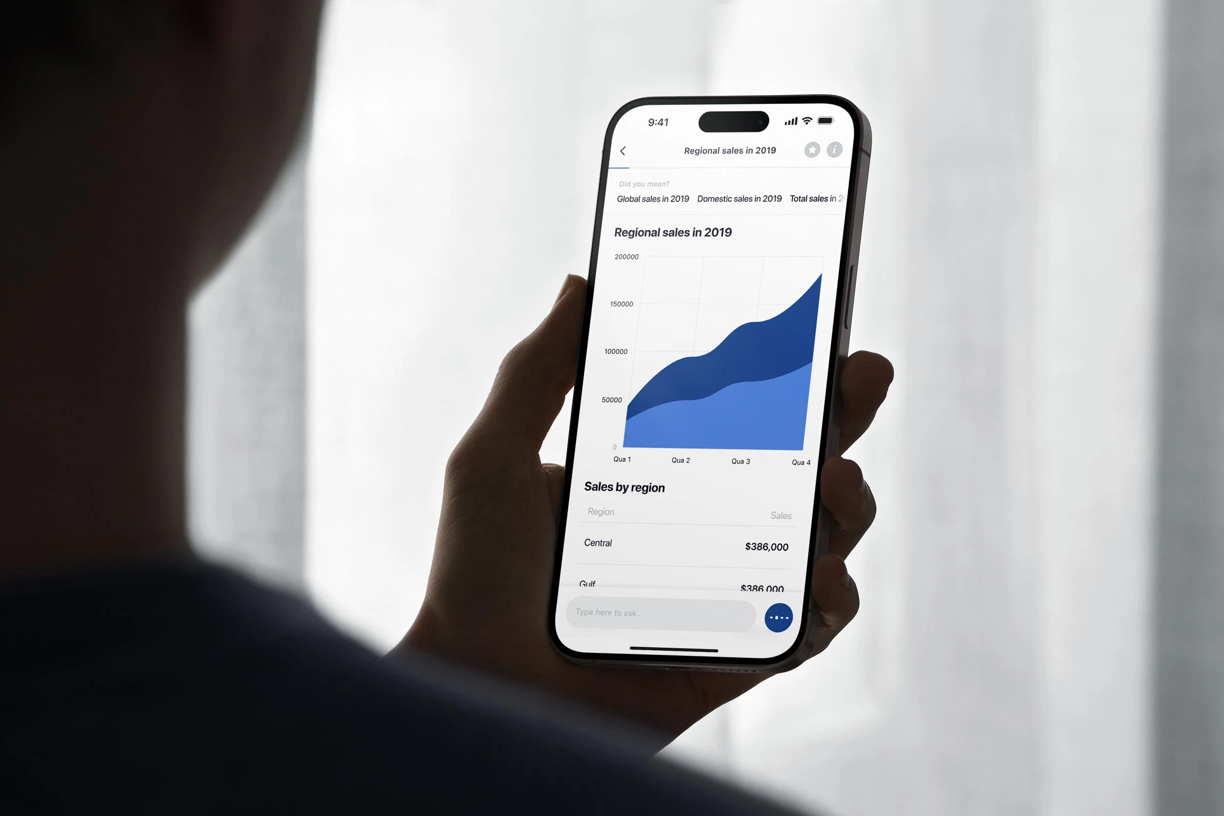 Stella answer view on an iPhone held by an out-of-focus figure. The query bar at top reads "Regional sales in 2019" with related-question chips beneath it (Global sales in 2019, Domestic sales in 2019, Total sales in 2019…). The answer is rendered as a stacked area chart, two-tone blue, with axes labelled 50000 to 200000 across Quarter 1 to Quarter 4. Below the chart, a "Sales by region" table begins (Central $386,000, Gulf $386,000…) with a "Type here to ask…" composer pinned to the bottom and the voice button beside it.